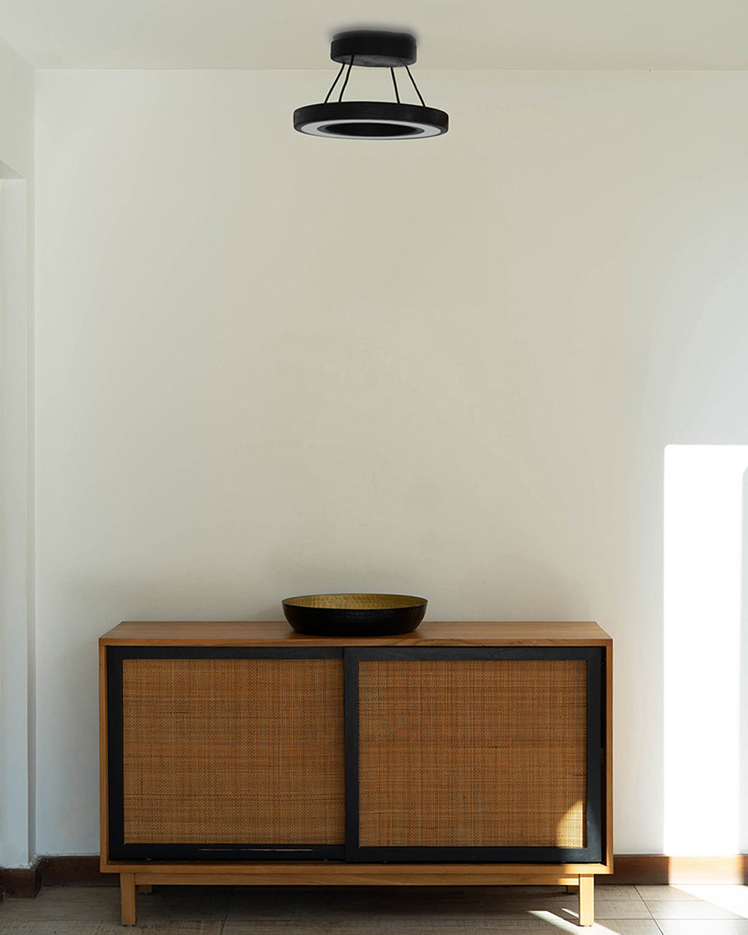 La escena muestra la lámpara de techo Runden en madera de pino con terminación en negro instalada en la entrada de una casa sobre un mueble de madera. Esta lámpara aporta al espacio haciéndolo moderno y elegante.