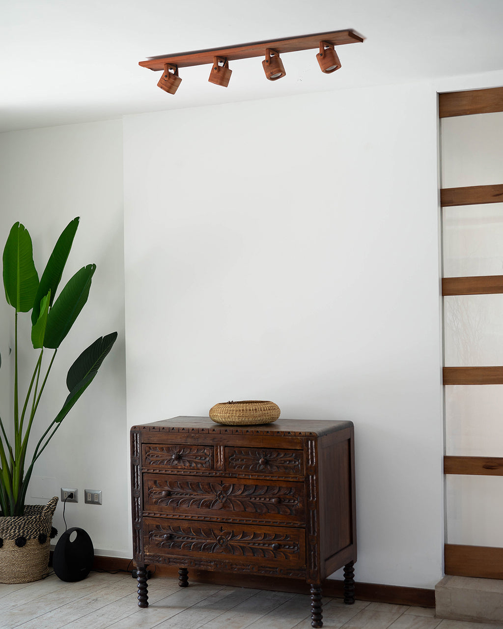 La escena muestra la aplicación del set de focos en madera de raulí en una sala donde se observa una planta, una cómoda y un separador de ambiente.