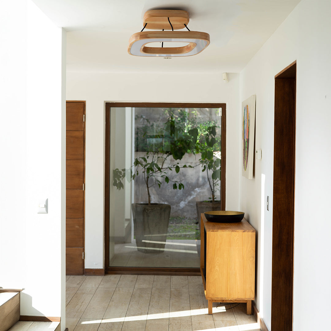 La escena muestra la lámpara de techo quadrat en madera de lenga instalada en la entrada de una casa con pasillo de madera. Esta lámpara aporta un toque natural y moderno al ambiente.