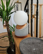 Lámpara de mesa con base de hormigón y madera. Pantalla con forma de esfera en vidrio blanco lechoso. Puesta en una mesa de entrada al lado de una planta.