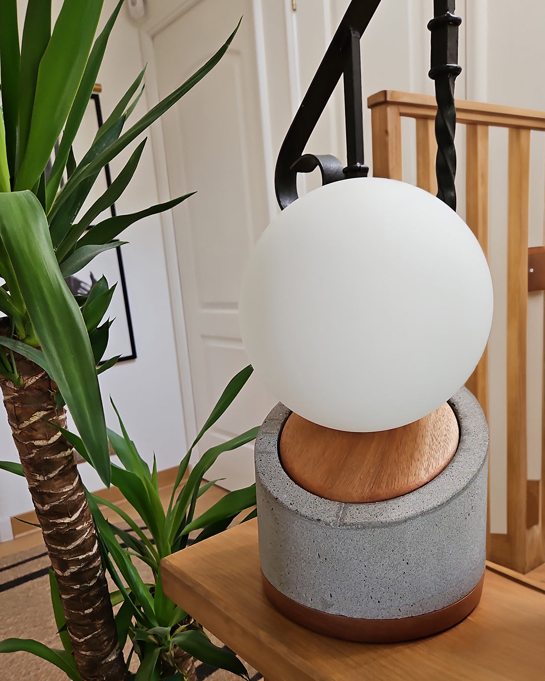 Lámpara de mesa con base de hormigón y madera. Pantalla con forma de esfera en vidrio blanco lechoso. Puesta en una mesa de entrada al lado de una planta.
