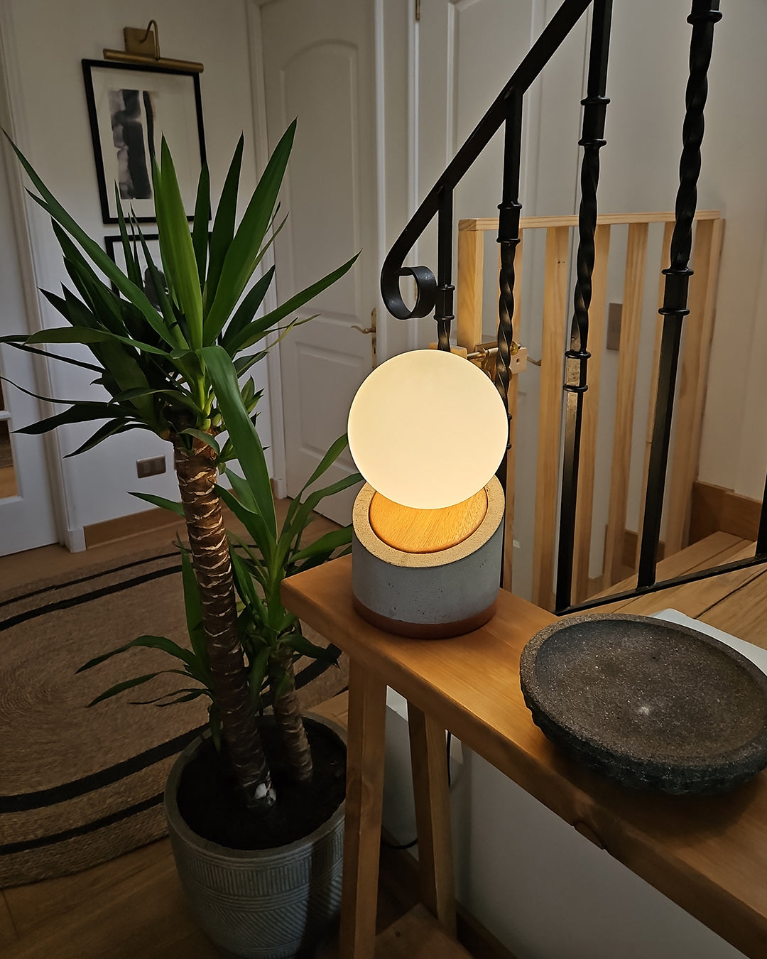 Lámpara de mesa con base de hormigón y madera. Pantalla con forma de esfera en vidrio blanco lechoso. Puesta en una mesa de entrada al lado de una planta y encendida.