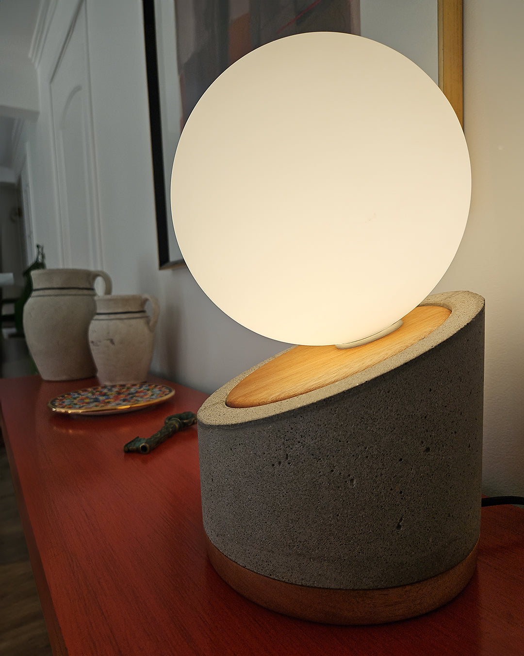 Lámpara de mesa con base de hormigón y madera. Pantalla con forma de esfera en vidrio blanco lechoso. Puesta en un rack rojo cerca de otros objetos decorativos. 