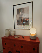 Lámpara de mesa con base de hormigón y madera. Pantalla con forma de esfera en vidrio blanco lechoso. Puesta en un rack rojo cerca de otros objetos decorativos.