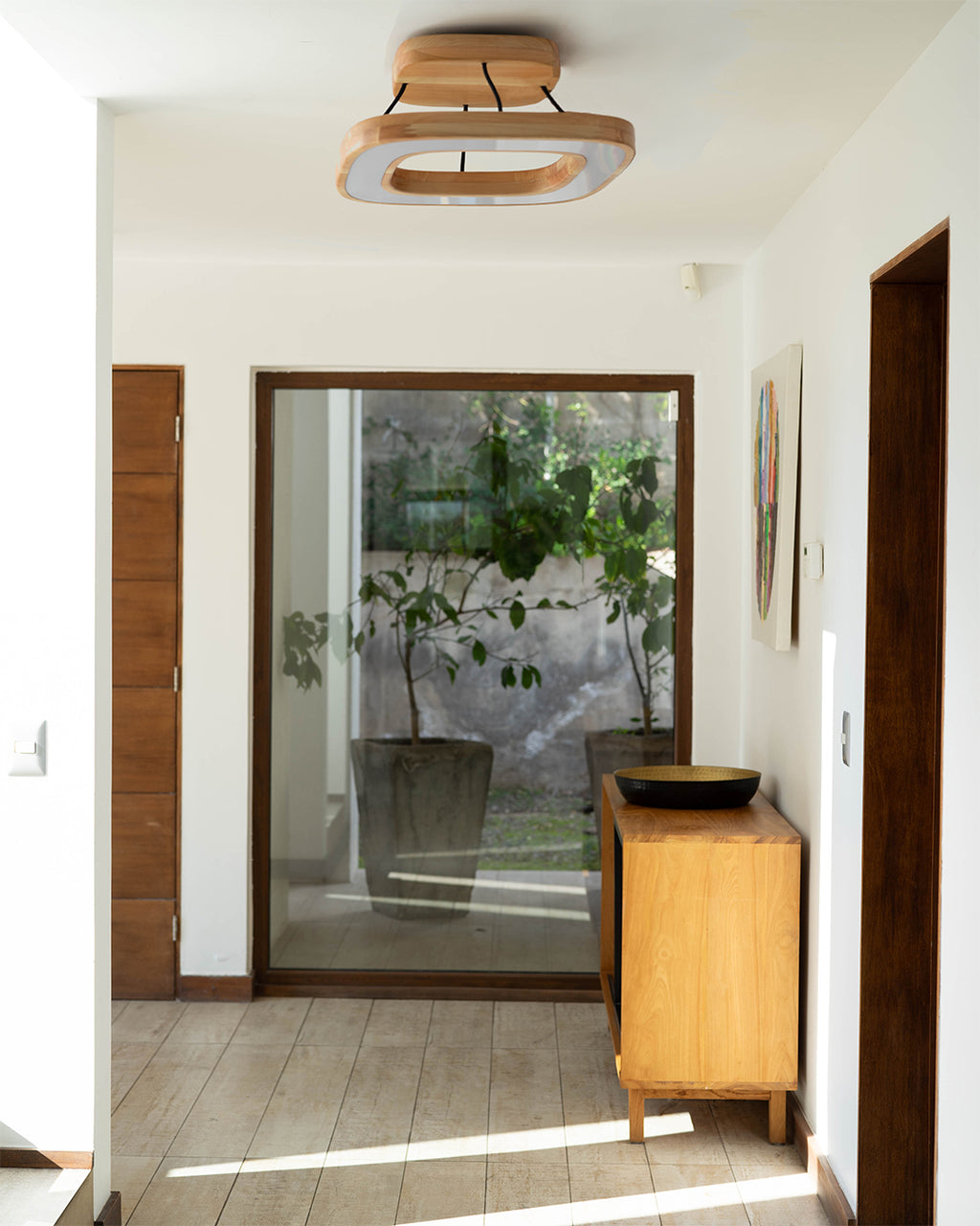 La escena muestra la lámpara de techo quadrat en madera de lenga instalada en la entrada de una casa con pasillo de madera. Esta lámpara aporta un toque natural y moderno al ambiente.