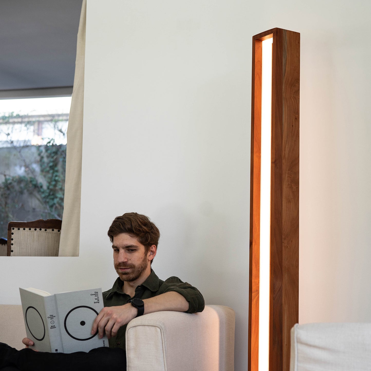 Hombre en un sofá, disfrutando de un libro, con la lámpara de pie caliza en madera de raulí iluminando el ambiente.